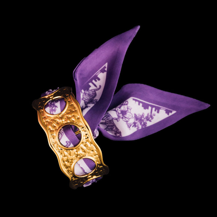 Gold cuff bracelets with purple scarf woven through it and tied in the back on a black background
