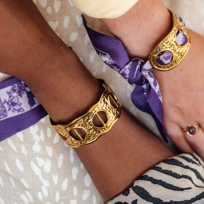 Close-up of hands wearing gold bracelets and a purple scarf with a lilac pattern pattern.