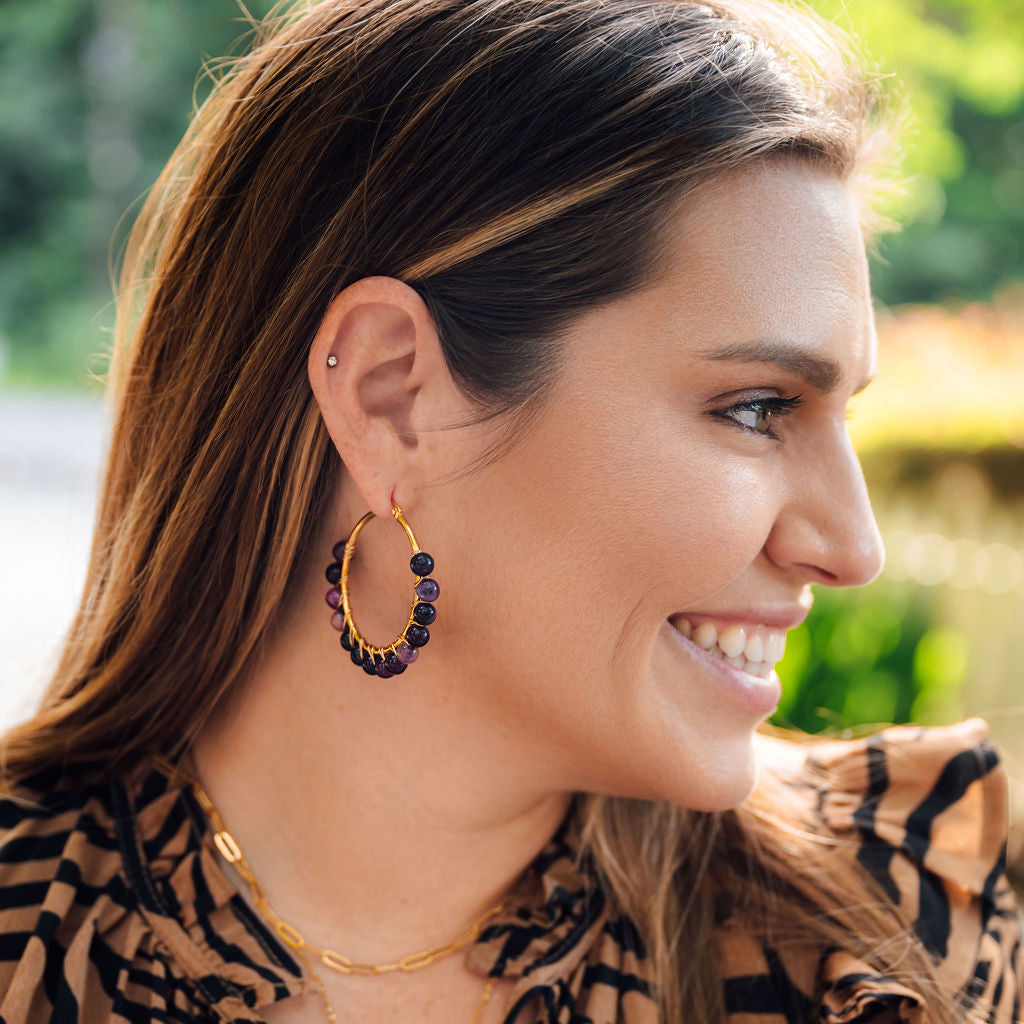Woman wearing amethyst hoop earrings and a tiger-striped blouse with a blurred outdoor background