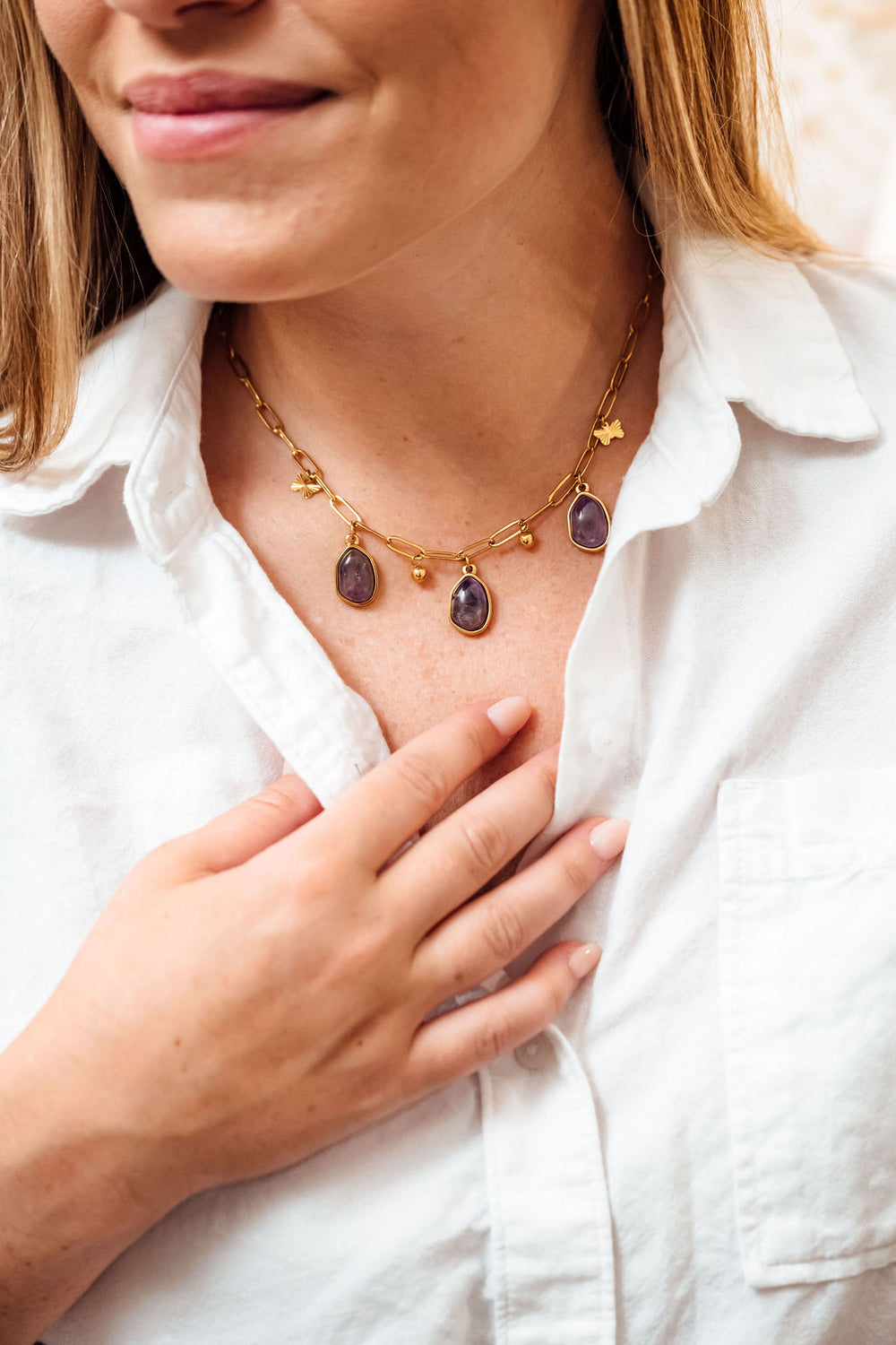 Woman wearing a gold necklace with purple stones, touching her chest.